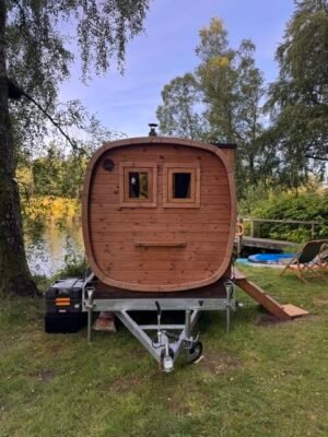 Vue avant du sauna mobile TimberIN Nordic Square sur remorque au bord d’un lac