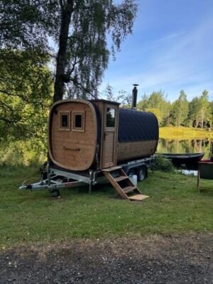 Vue latérale du sauna mobile TimberIN Nordic Square sur remorque avec cheminée au bord d’un lac
