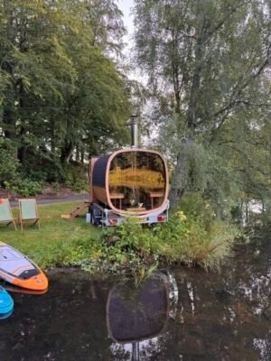 Vue arrière panoramique vitrée du sauna mobile TimberIN Nordic Square sur remorque au bord d’un lac