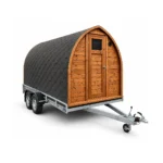 Sauna extérieur 15 Sauna mobile en forme de pod TimberIN Nordic, avec façade en bois, porte pleine, coque en bardeaux noirs et remorque à un essieu sur fond blanc