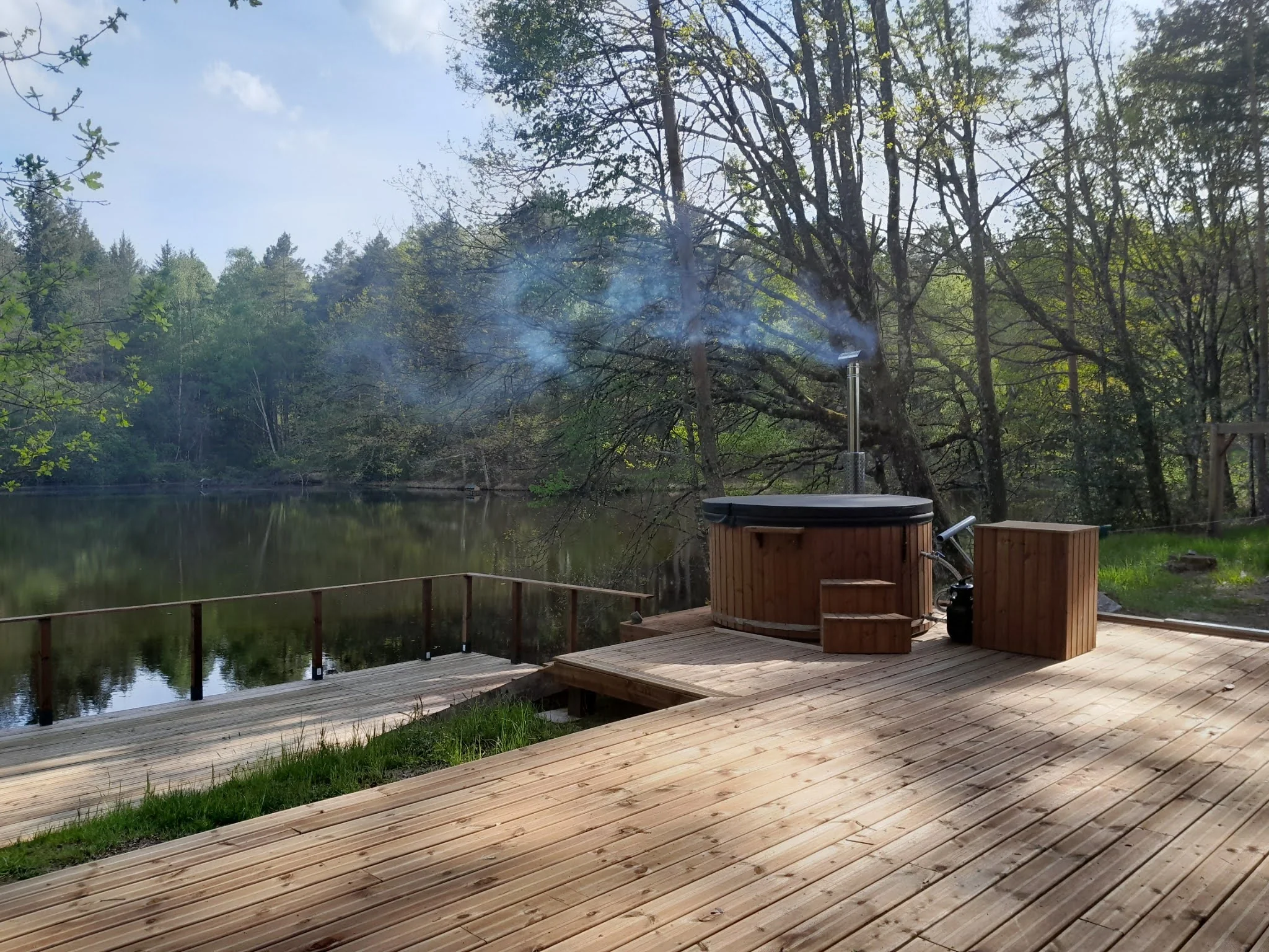 Pourquoi choisir TimberIN ? 5 Bain nordique en bois avec cheminée en fonctionnement sur une terrasse au bord d’un lac entouré d’arbres