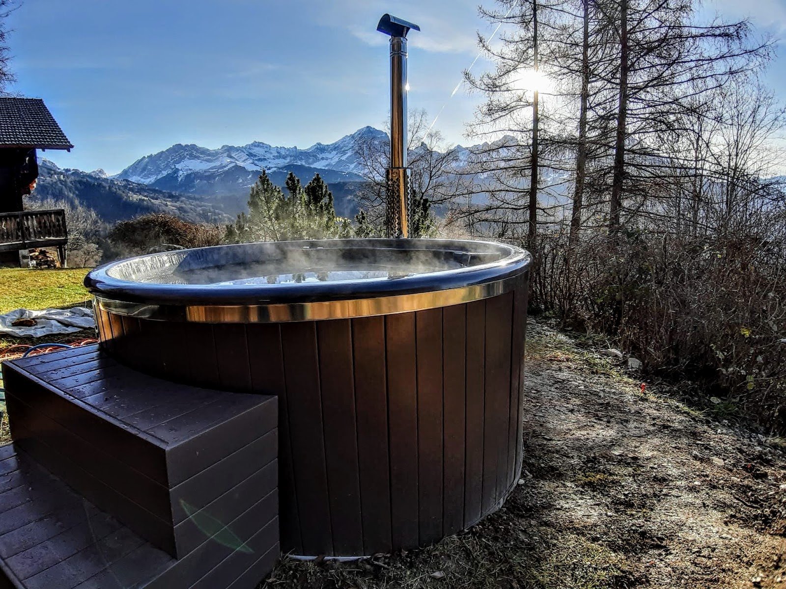 Home 1 Bain nordique rond fumant avec poêle extérieur et cheminée inox devant des montagnes enneigées
