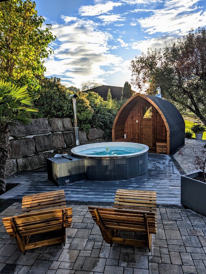 Exigences électriques 1 Bain nordique et sauna en bois aménagés sur une terrasse de jardin bordée de pierres et de végétation